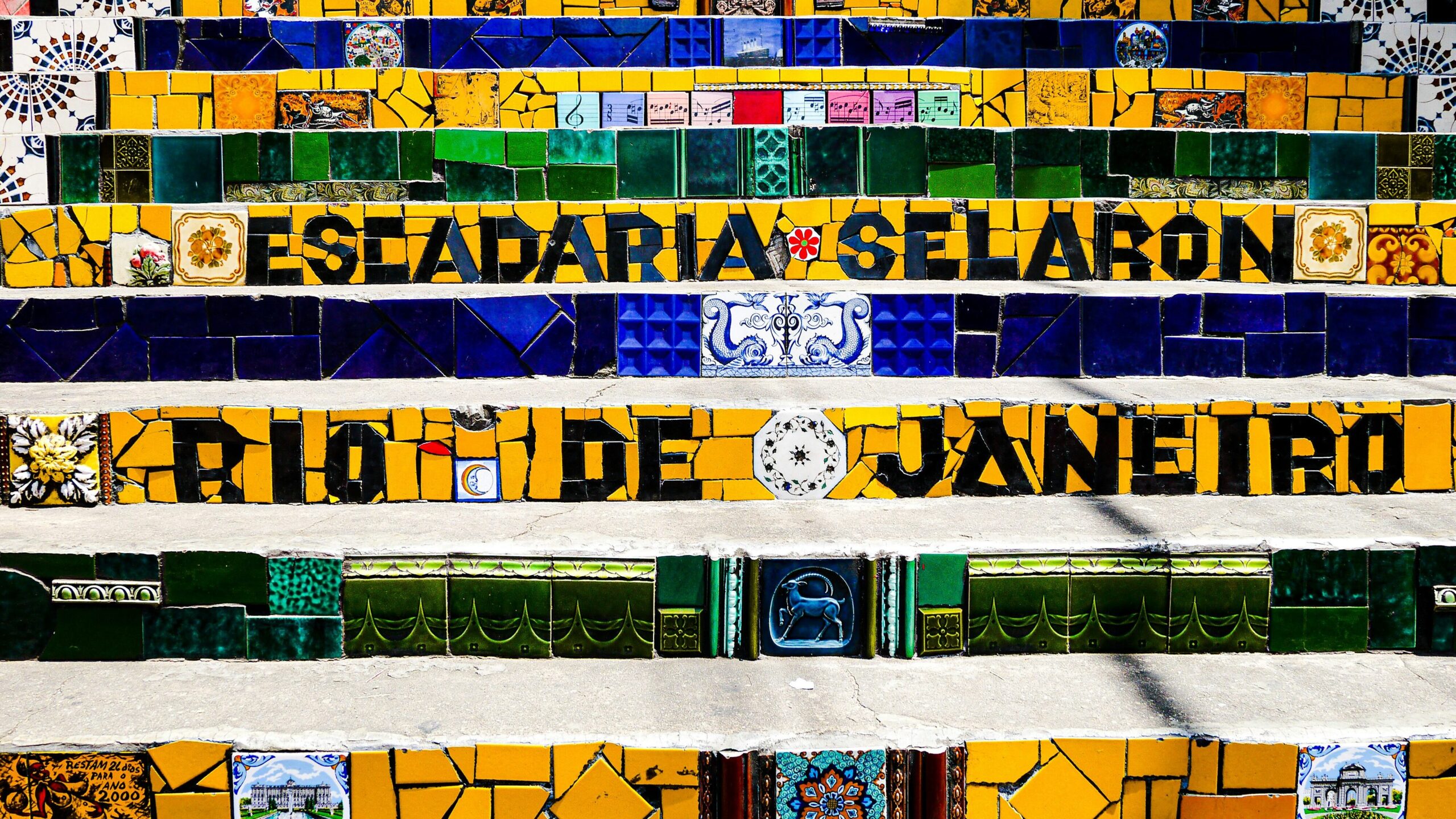 Close-up view of the Selarón Stairs in Rio de Janeiro, focusing on a few vibrant, colorful tiles. The tiles prominently feature the inscription 'Escadaria Selarón Rio de Janeiro' in bold, artistic lettering, surrounded by intricate patterns and bright, lively colors that reflect the iconic mosaic style of the staircase.