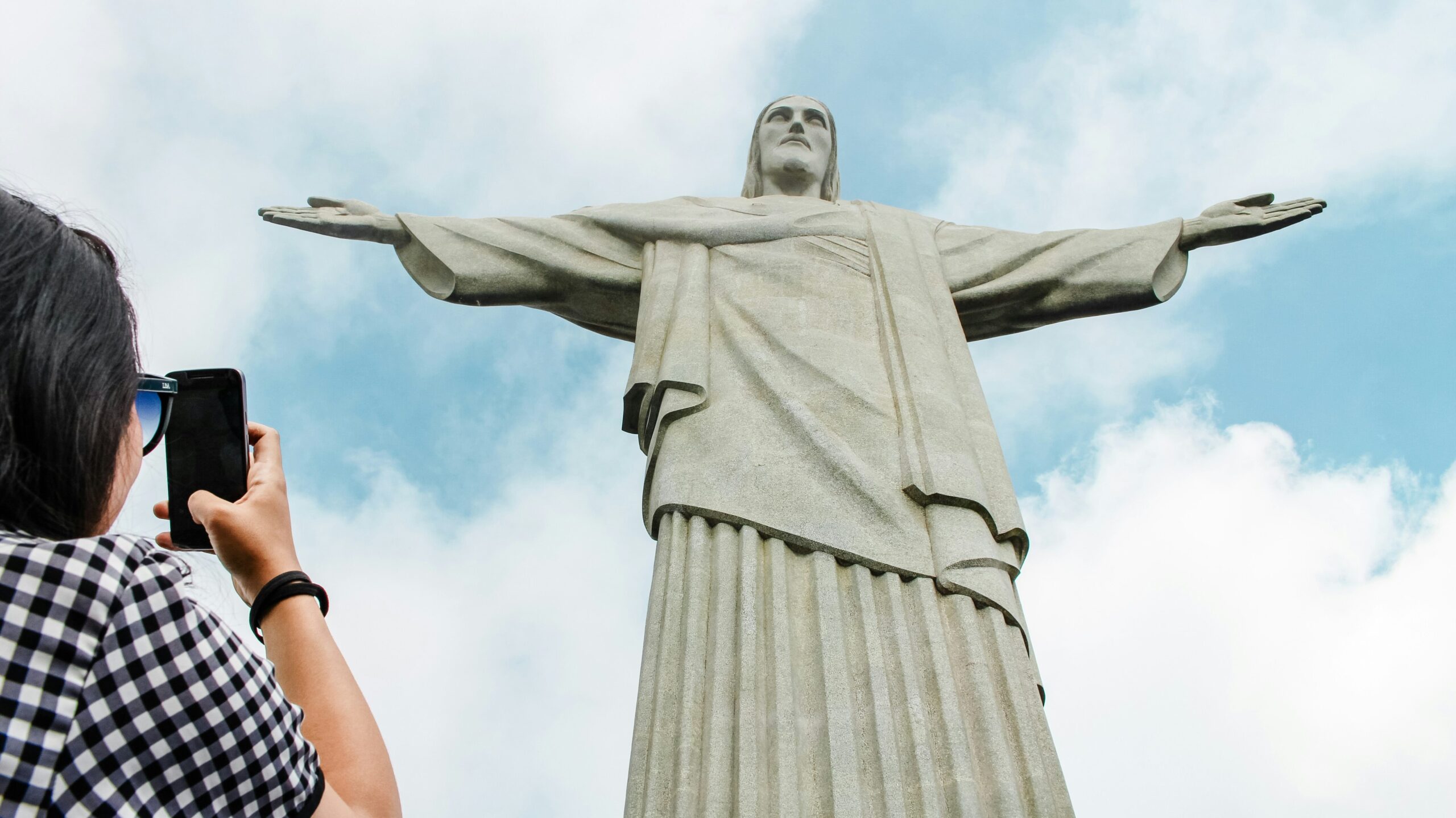 A person taking a photo of the iconic Christ the Redeemer statue in Rio de Janeiro against a backdrop of blue sky and clouds.