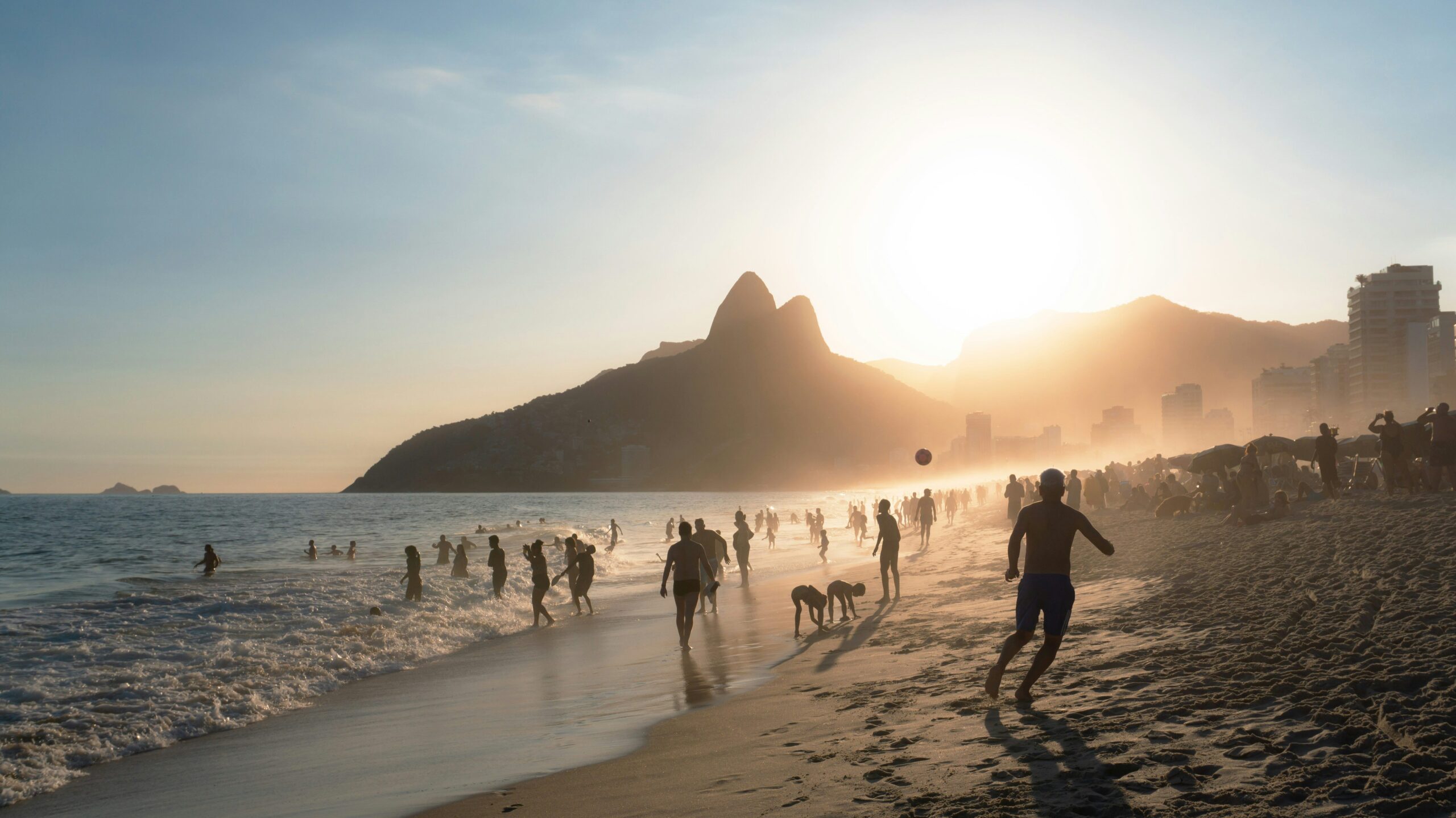 A vibrant scene at Ipanema Beach in Rio de Janeiro during sunset, with people enjoying the water, playing beach games, and relaxing against the backdrop of the iconic Two Brothers Mountains.