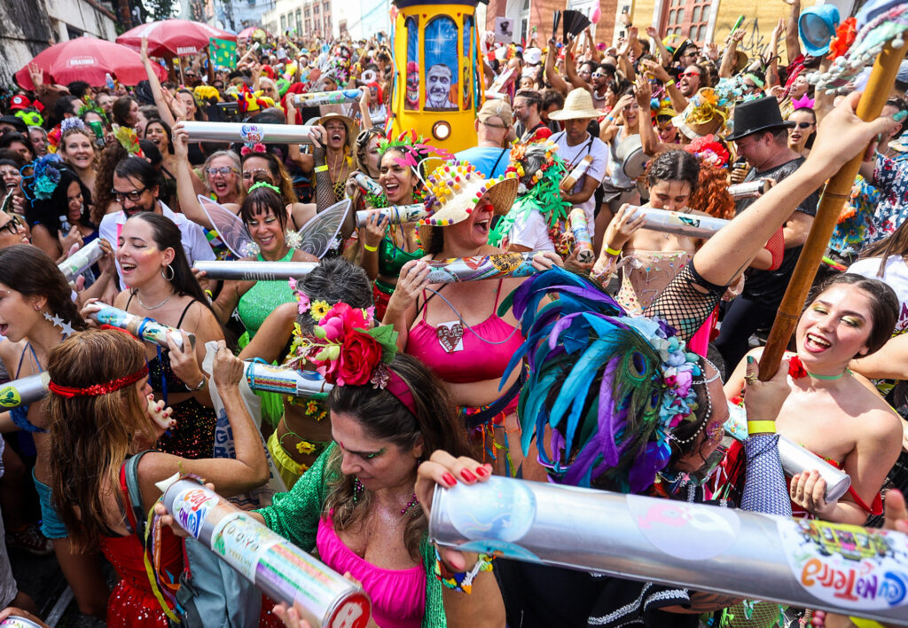 A lively street party during Rio de Janeiro’s Carnival, featuring a diverse crowd dressed in colorful and extravagant costumes adorned with flowers, feathers, and glitter. Participants dance joyfully, holding and shaking large metallic tubes, while a small, decorated yellow tram is visible in the background. The streets are packed with revelers celebrating under the bright sunlight, capturing the energetic and festive spirit of Brazil’s famous Carnival blocos (street parties).