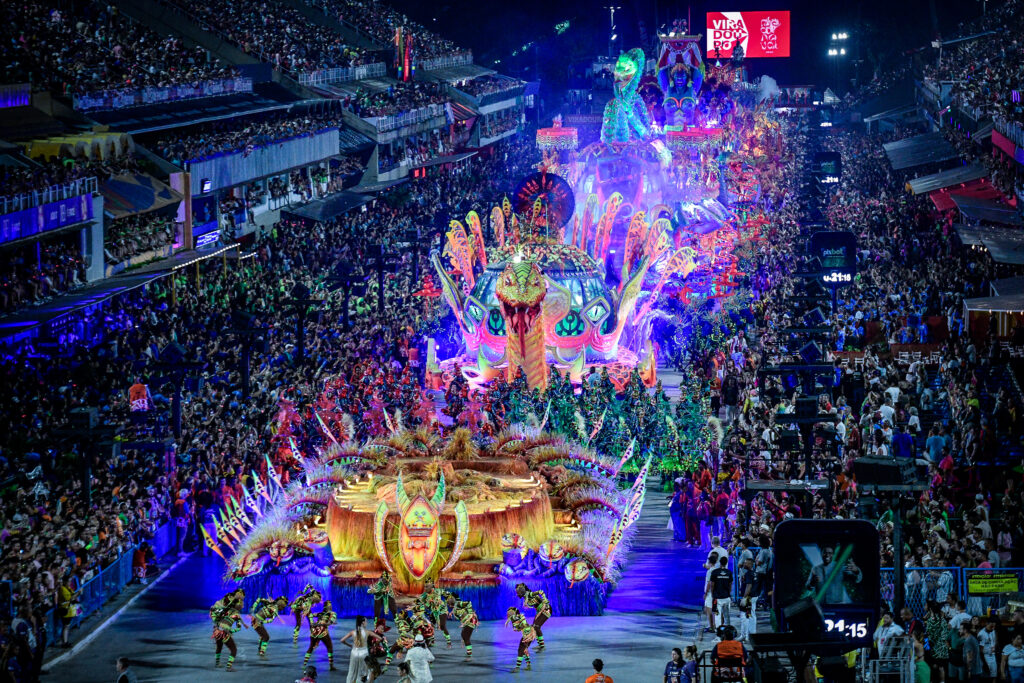 A vibrant scene from Rio de Janeiro's Carnival parade at the Sambadrome, featuring a massive, colorful float with a serpent-like design, glowing neon lights, and intricate decorations. Dancers in elaborate costumes perform energetically in front of the float, while grandstands packed with spectators cheer and watch the spectacle. The atmosphere is electric with bright lights, music, and movement, showcasing the grandeur of Brazil’s famous festival.